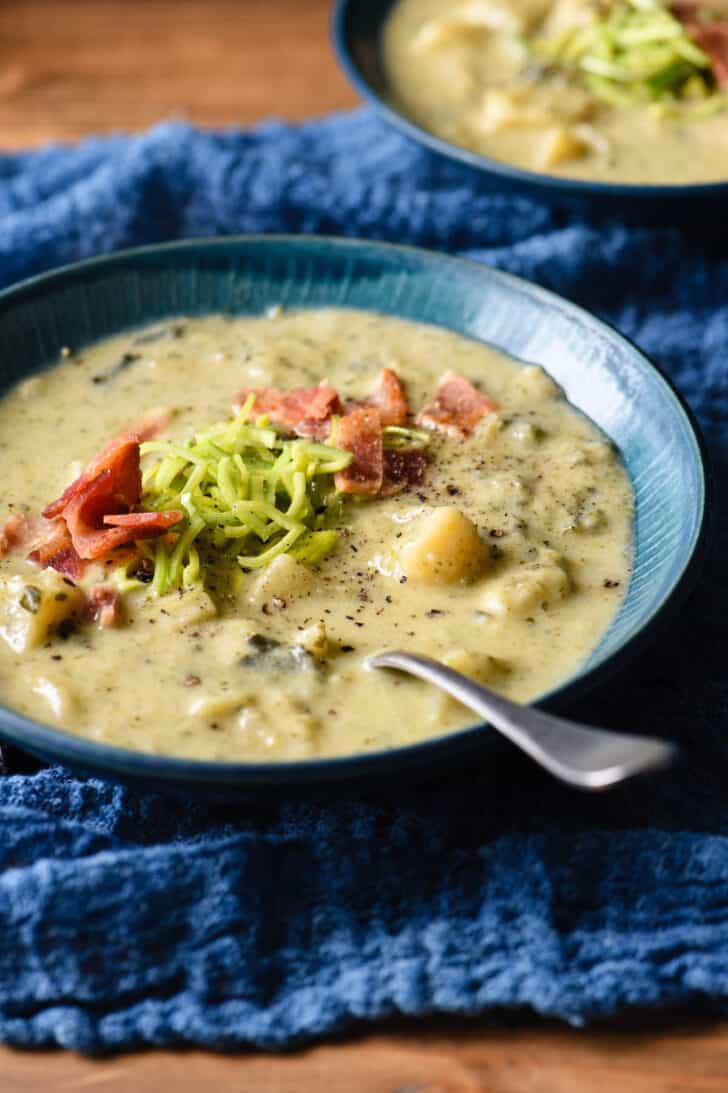 A blue bowl filled with colcannon soup garnished with sliced leeks and bacon, with a spoon digging into it, on top of a blue napkin on a wooden table.