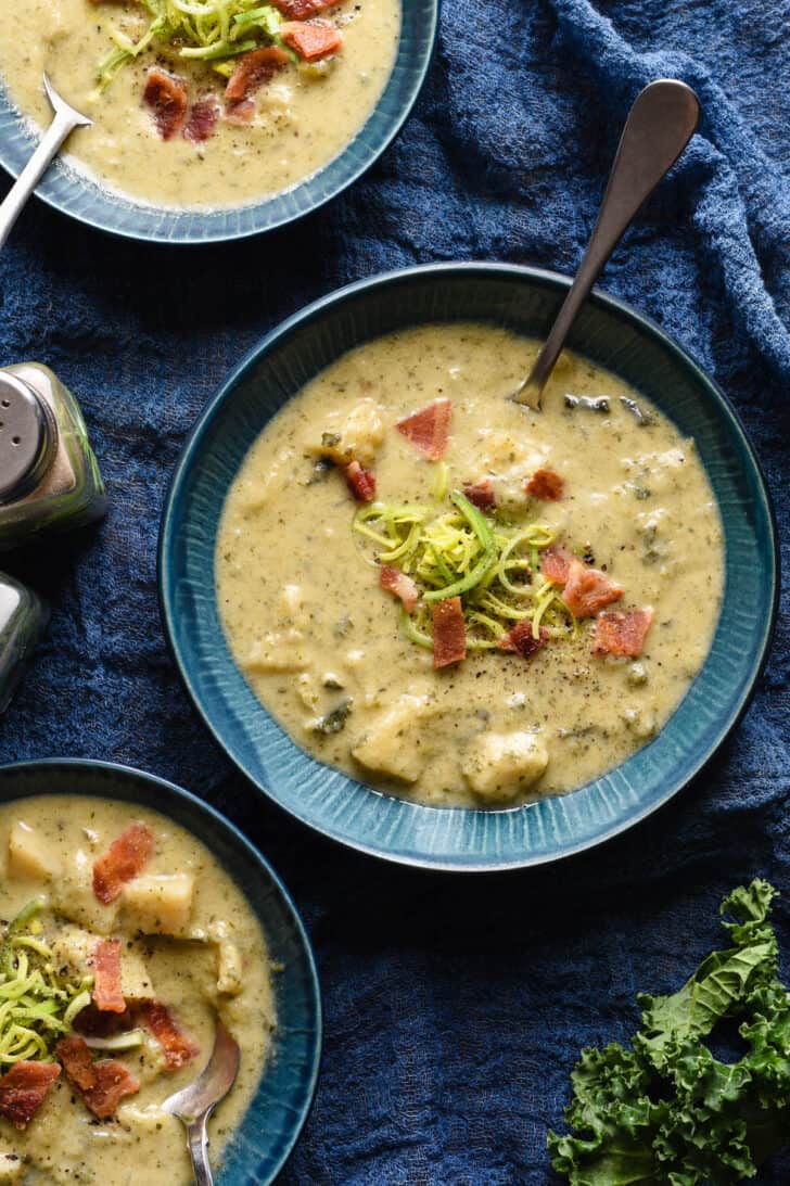 Three blue bowls on a blue linen, filled with cabbage potato soup garnished with sliced leeks and crispy bacon.