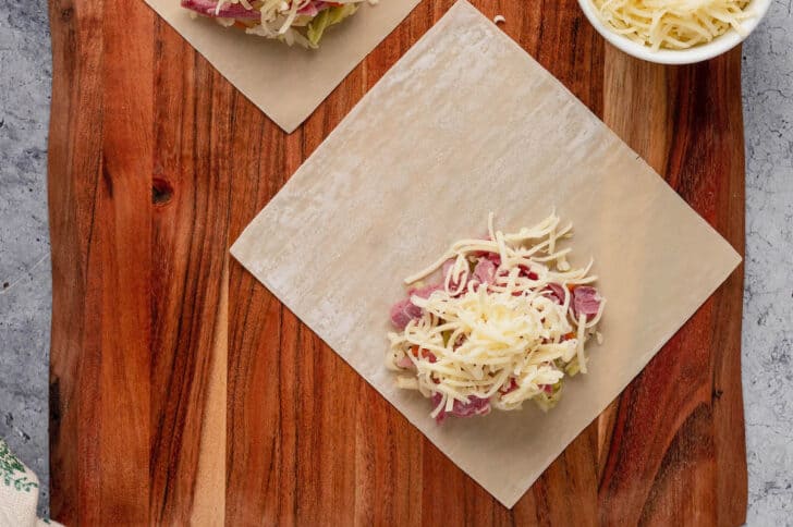An egg roll wrapper on a wooden cutting board with a dollop of coleslaw, corned beef and shredded white cheese on it.
