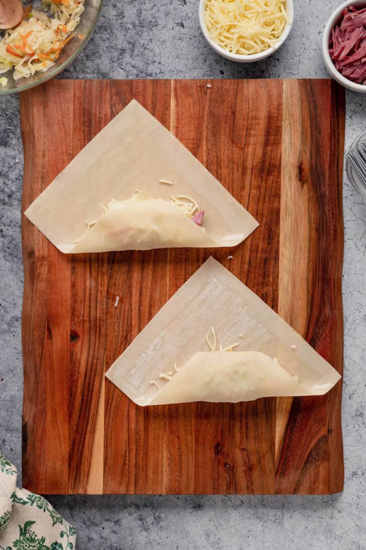 Corned beef egg rolls being rolled up on a wooden cutting board.