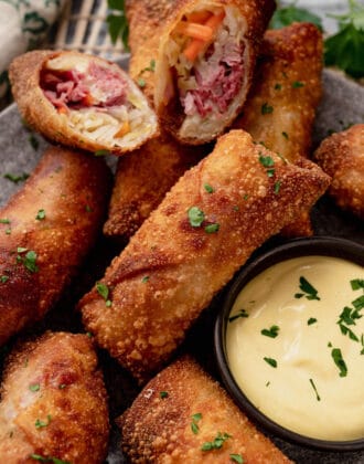 A plate of corned beef egg rolls with creamy mustard dipping sauce. One egg roll is cut in half, showing the inside filling.