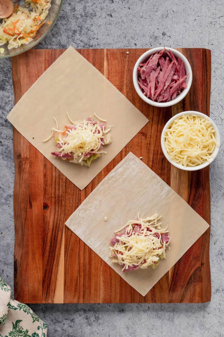 Egg roll wrappers laid out on a wooden cutting board, topped with small pies of corned beef and shredded cheese.