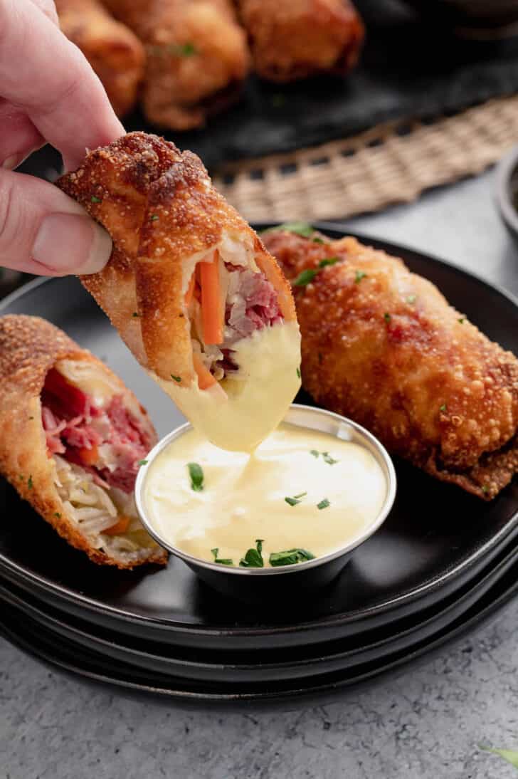 A half of a reuben egg roll being dipped into a creamy mustard sauce.