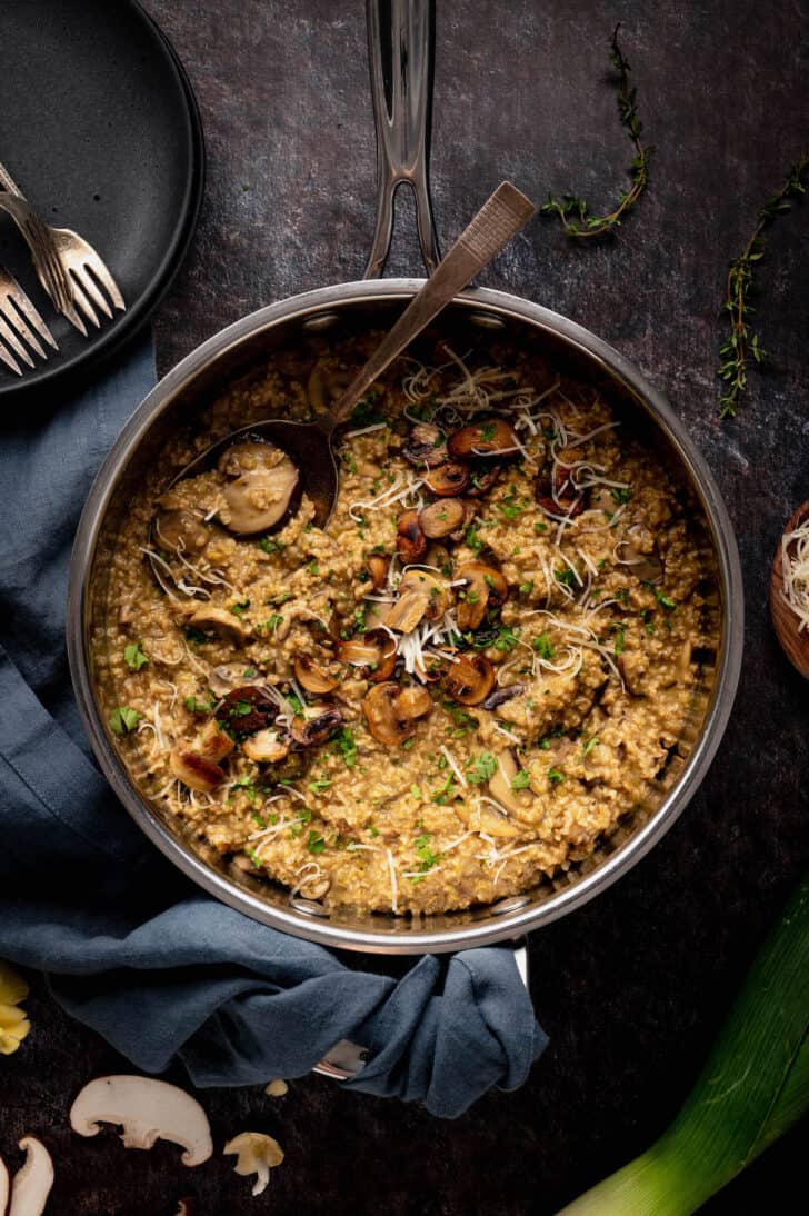 A stainless steel skillet filled with savory oatmeal with mushrooms and cheese, with a serving spoon digging into it.