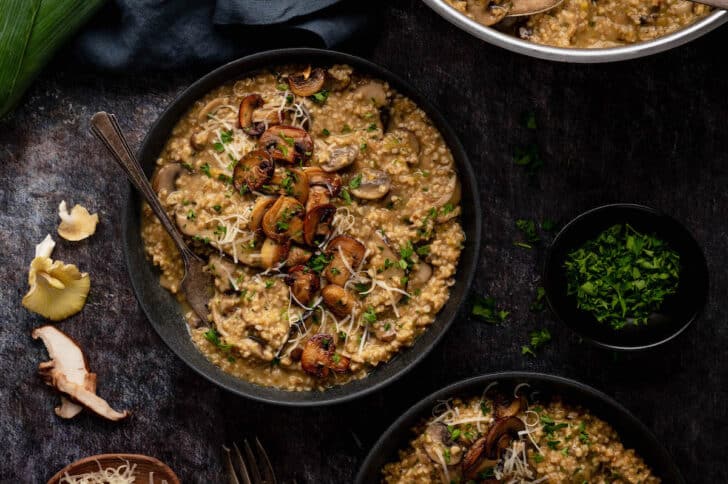 Large dark shallow bowls filled with oat risotto topped with mushrooms on a dark surface.