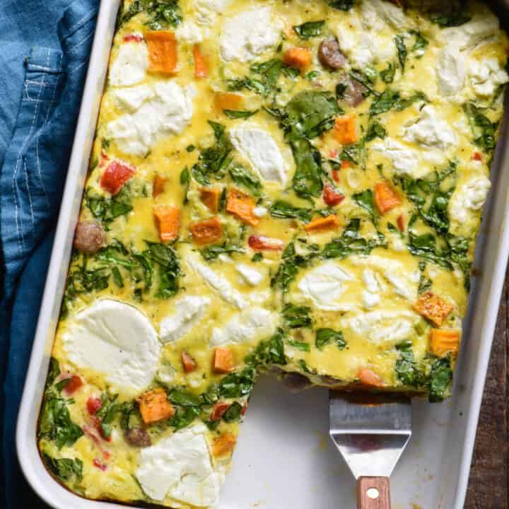 Sweet potato breakfast casserole made with eggs, sausage, spinach and goat cheese, in rectangular white baking dish, with a serving spatula lifting a piece out.