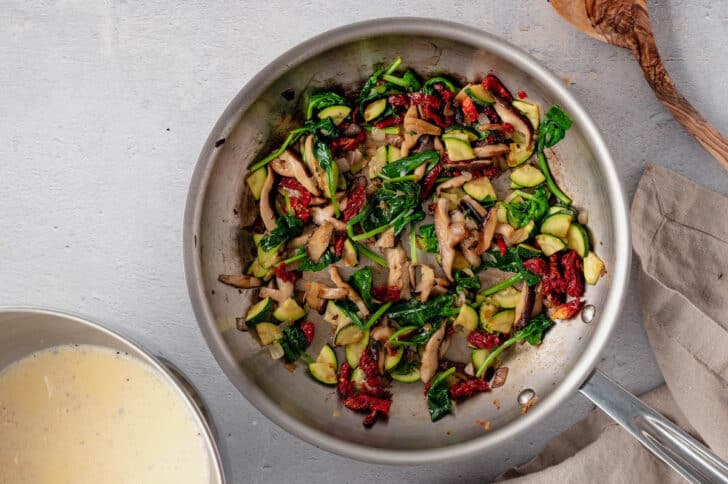 Mushrooms, shallots, zucchini and spinach being sauteed in a stainless steel skillet for a vegetable quiche.