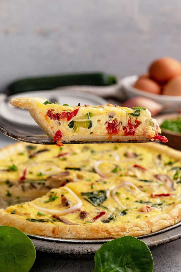 A slice of veggie quiche being lifted out of a full quiche in a metal pie plate.