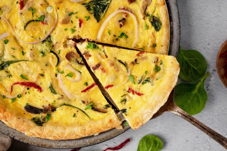 A slice of vegetable quiche being lifted out of a full quiche in a metal pie plate.