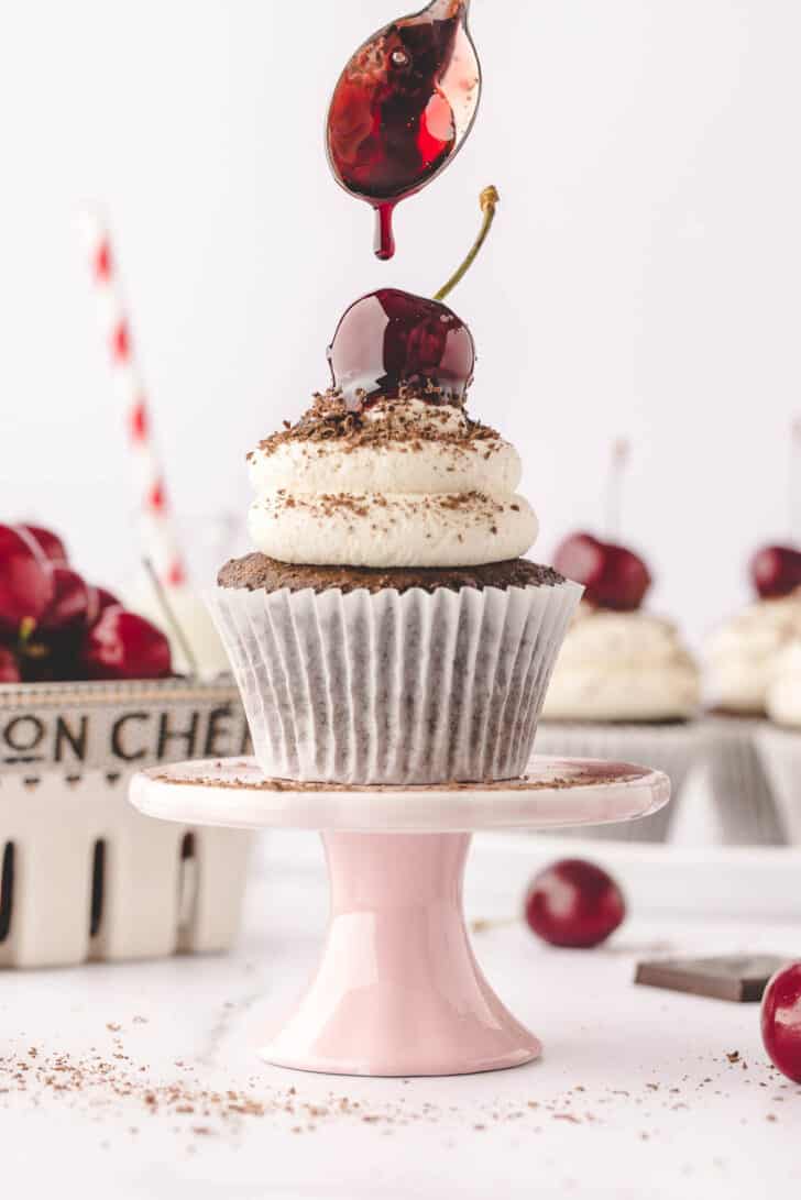 A chocolate cupcake topped with whipped cream, chocolate shavings and a cherry, on a small pink cake stand, with a spoon drizzling cherry syrup over it.