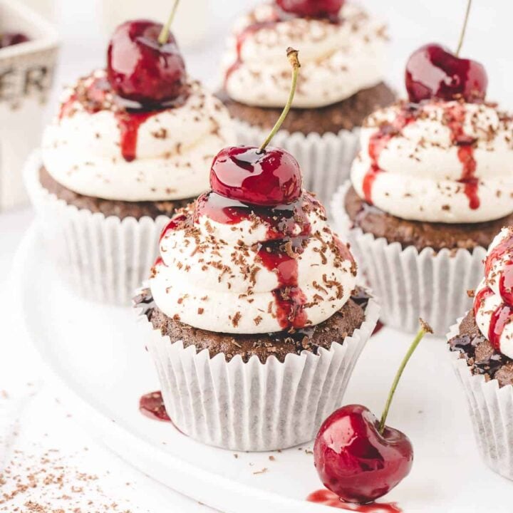 A white plate of Black Forest cupcakes, which are chocolate cupcakes topped with whipped cream, cherries and chocolate shavings.