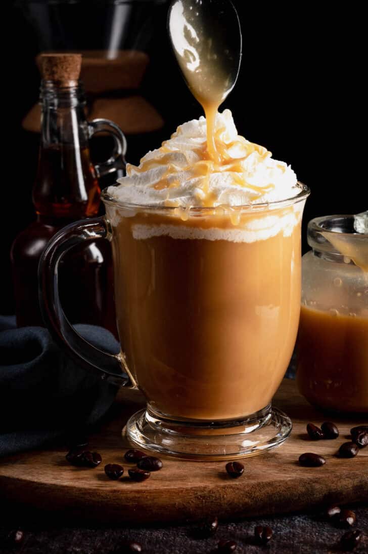 A glass mug of java topped with whipped cream and being drizzled with a brown dessert sauce.