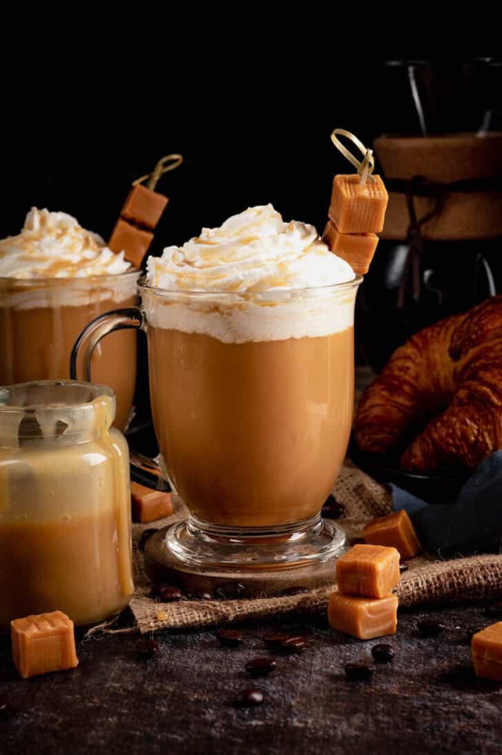 A glass mug filled wth creamy caramel coffee and garnished with whipped cream and a skewer of caramel candies.