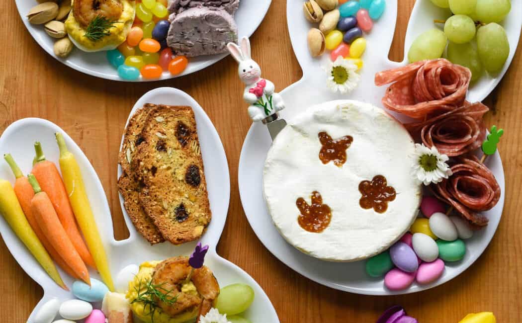 An Easter charcuterie board arranged on three white bunny head shaped plates. The plates are filled with charcuterie, cheeses, crackers, fruit, carrots, and candy.