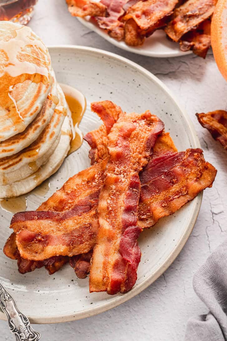 A speckled plate filled with a pile of bacon slices and a stack of pancakes with syrup.