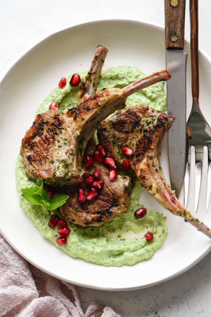 Lollipop lamb chops garnished with pomegranate seeds on top of a creamy green sauce, in a white shallow bowl.