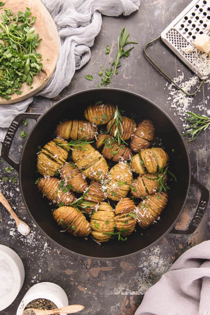 Mini Hasselback potatoes in a cast iron skillet, sprinkled with rosemary sprigs and shredded Parmesan cheese.