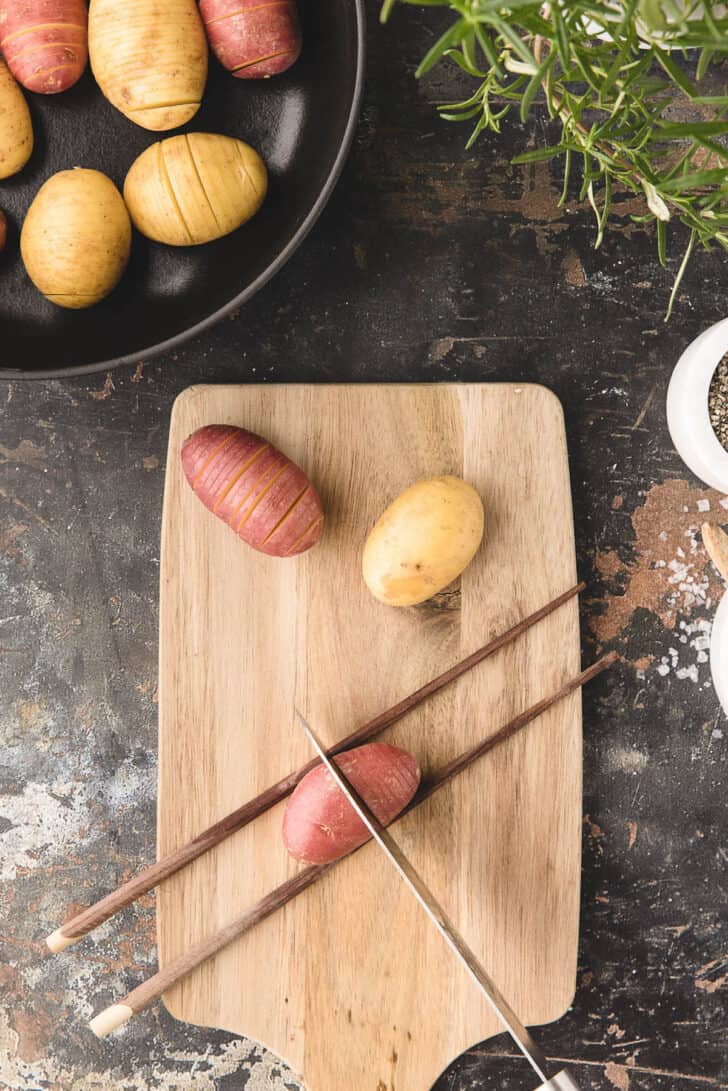 A small red potato between two chopsticks on a cutting board, with a knife slicing it crosswise.