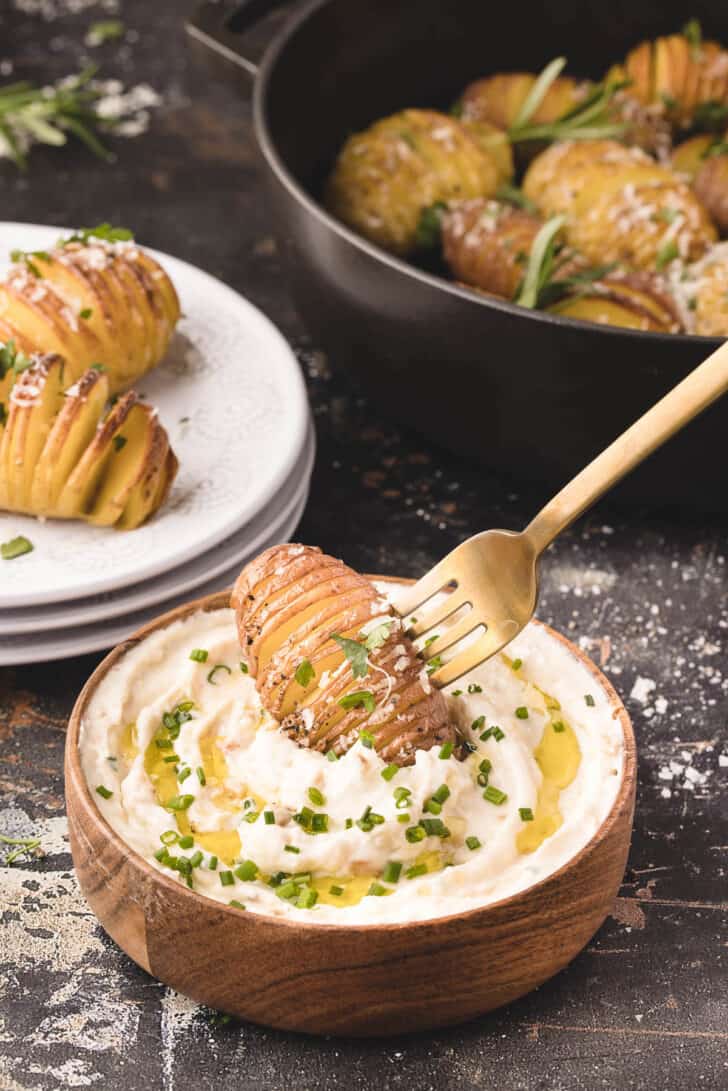 A gold fork dipping a mini Hasselback potato into a wooden bowl filled with a creamy dipping sauce topped with olive oil and chives.