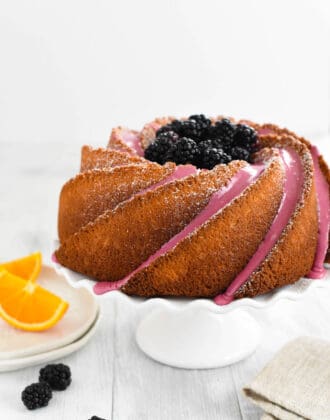 An orange bundt cake decorated with blackberry icing and fresh blackberries, on a white fluted cake stand.