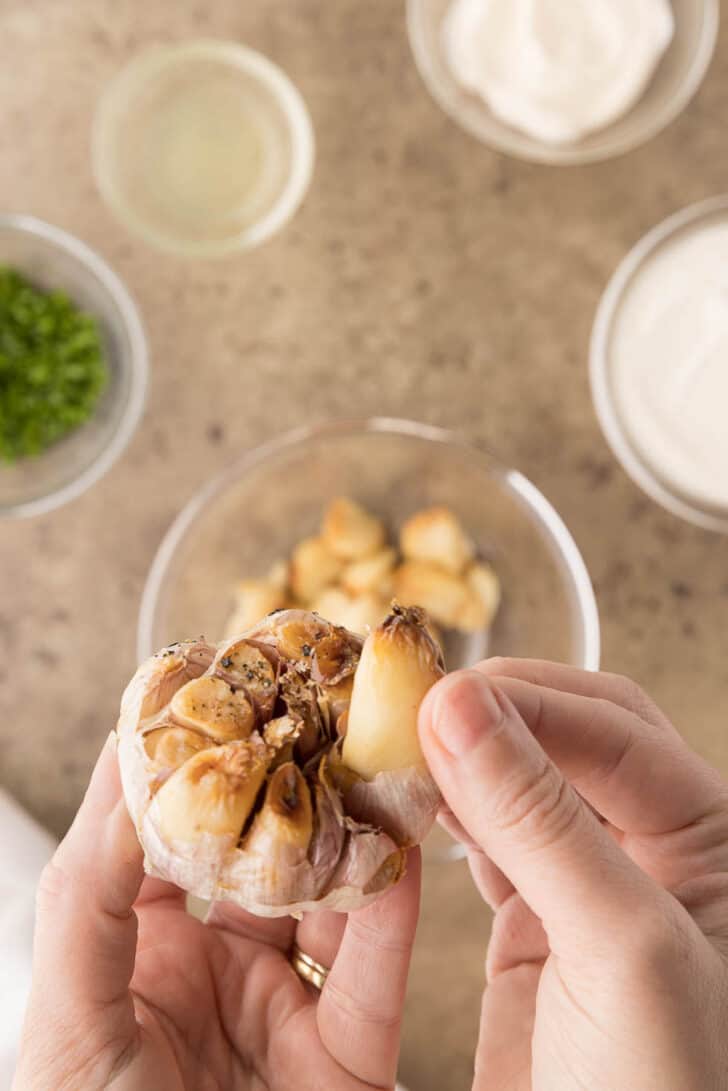 Hands holding a roasted head of garlic and removing one of the cloves from the head, over a bowl.