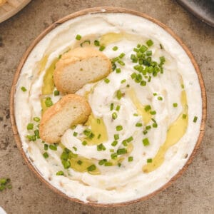 A round bowl filled with creamy roasted garlic dip garnished with chives and toasted bread slices.