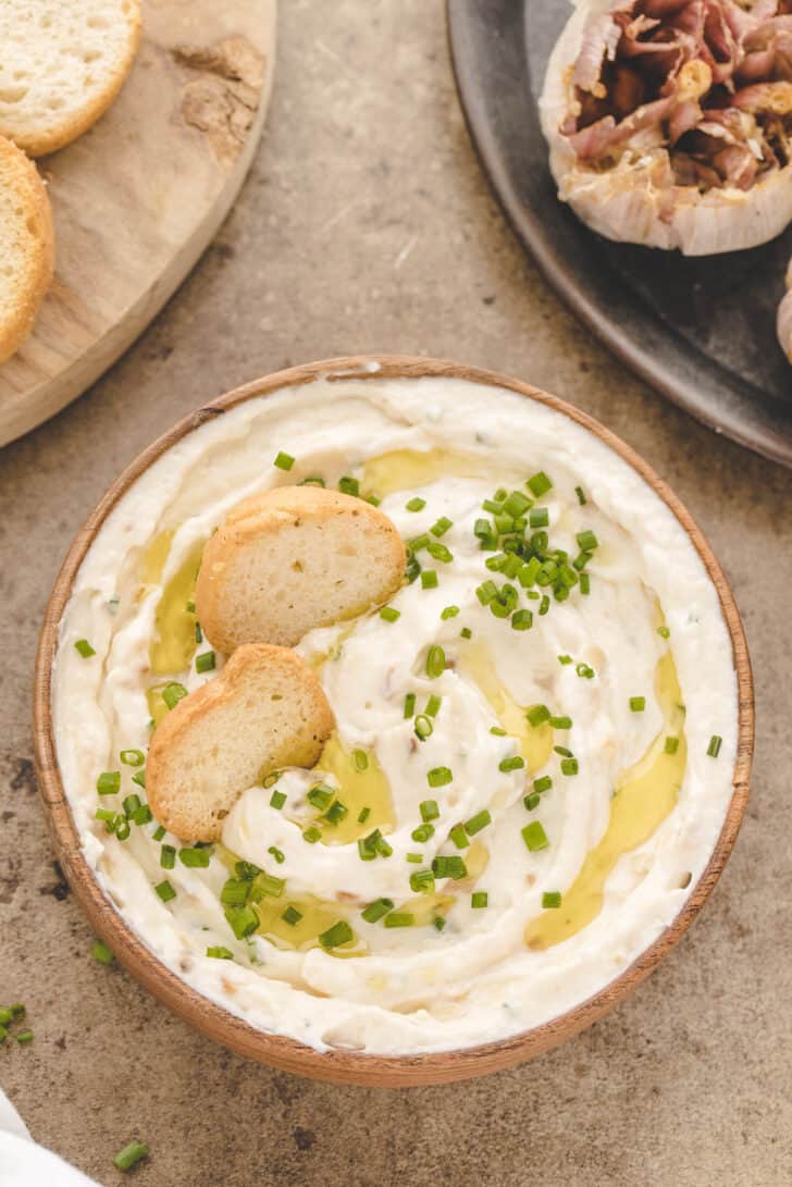 A round bowl filled with creamy roasted garlic dip garnished with chives and toasted bread slices.