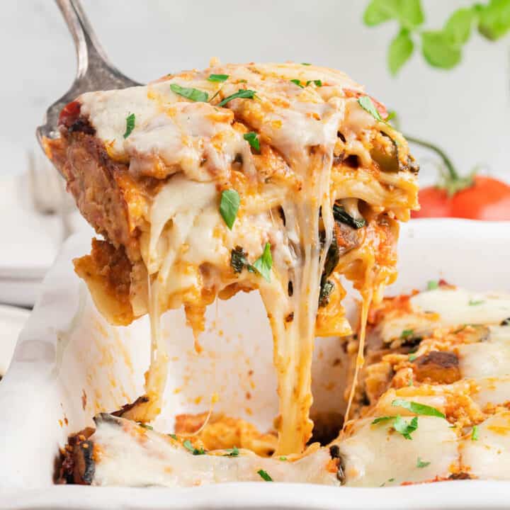 A piece of vegetable lasagna garnished with small basil leaves being lifted out of a white baking dish, with gooey cheese stretching between the piece and the dish.
