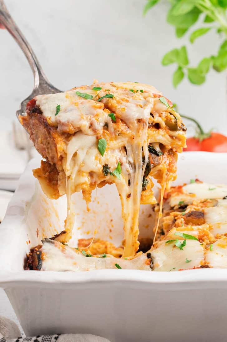 A piece of vegetable lasagna garnished with small basil leaves being lifted out of a white baking dish, with gooey cheese stretching between the piece and the dish.