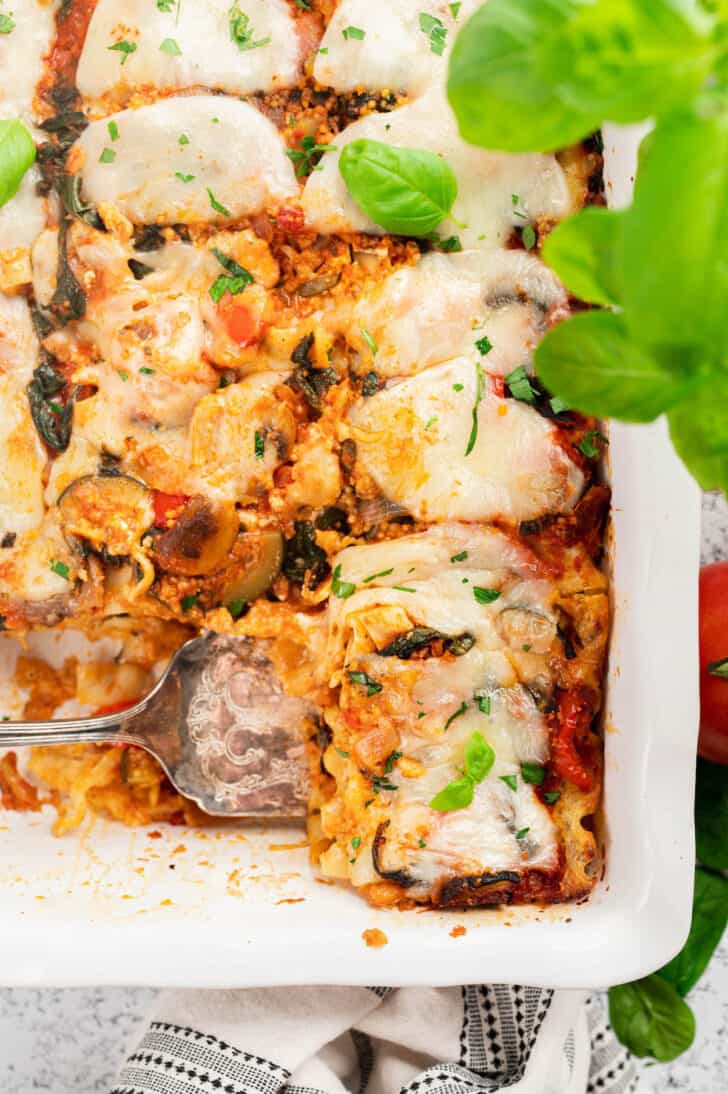 A white baking dish filled with a veggie lasagna recipe, with a serving utensil about to lift one piece out.