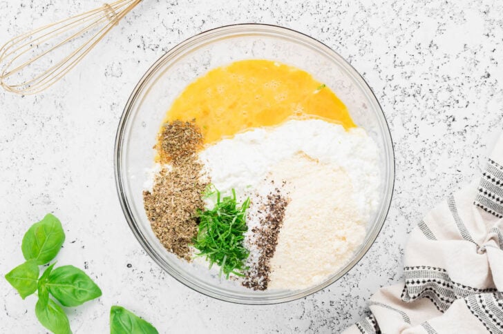 A glass bowl filled with cottage cheese, Parmesan cheese, a beaten eggs, herbs and spices.