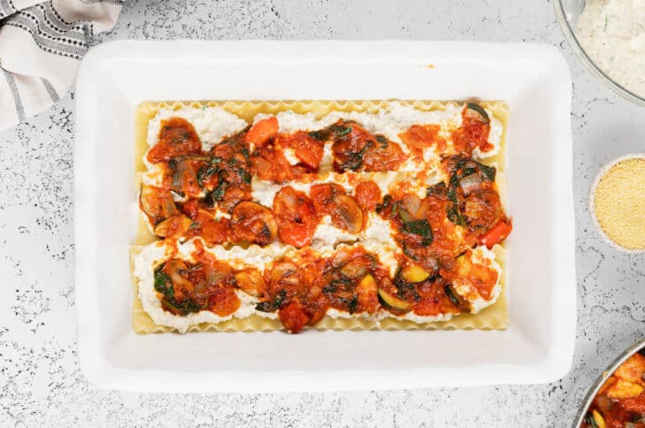 A white lasagna dish layered with lasagna noodles, a cheese mixture and a vegetable marinara mixture.
