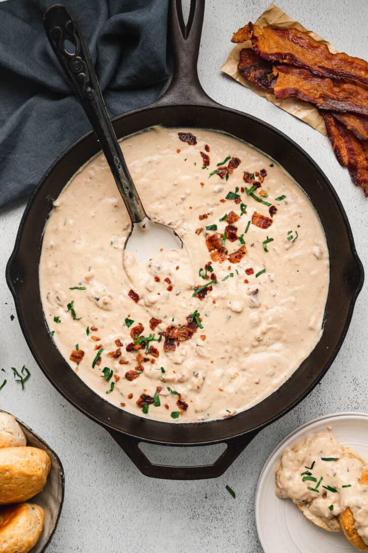 A cast iron skillet full of bacon gravy, garnished with chopped bacon and green herbs, with a spoon digging into it.