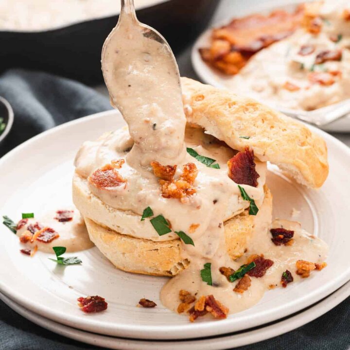 A biscuit cut in half on a small white plate, with bacon gravy being spooned over it.