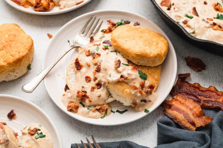 A plate of biscuits and bacon gravy, garnished with chopped bacon and green herbs.