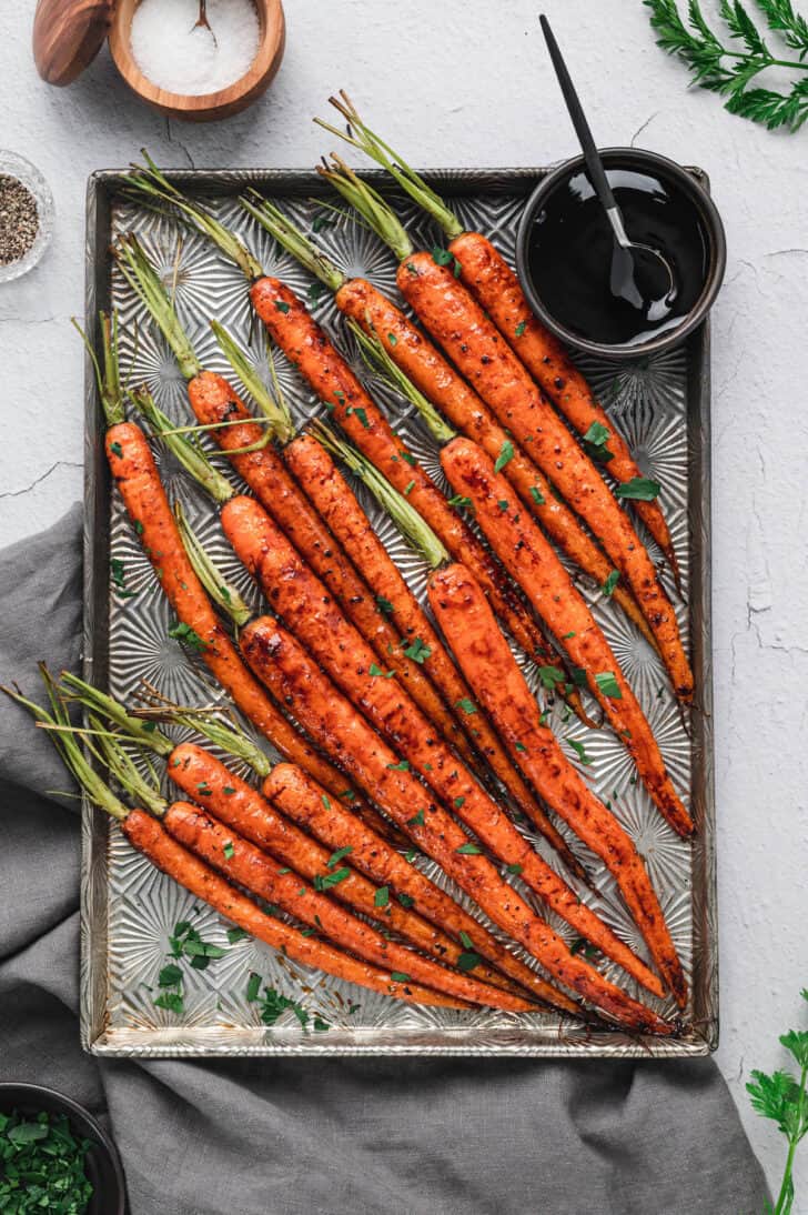 A textured baking pan topped with balsamic roasted carrots with their green tops still on, with a small bowl of balsamic glaze on the pan.