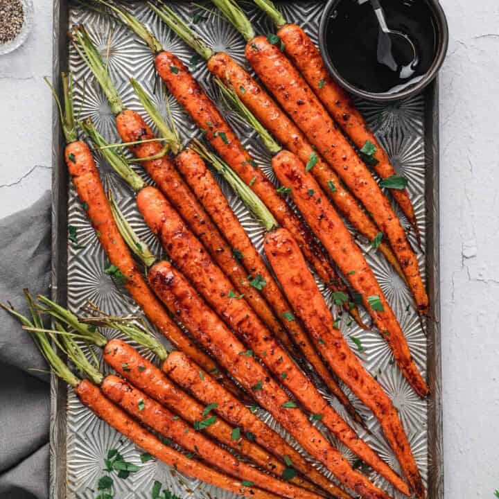 A textured baking pan topped with balsamic roasted carrots with their green tops still on, with a small bowl of balsamic glaze on the pan.