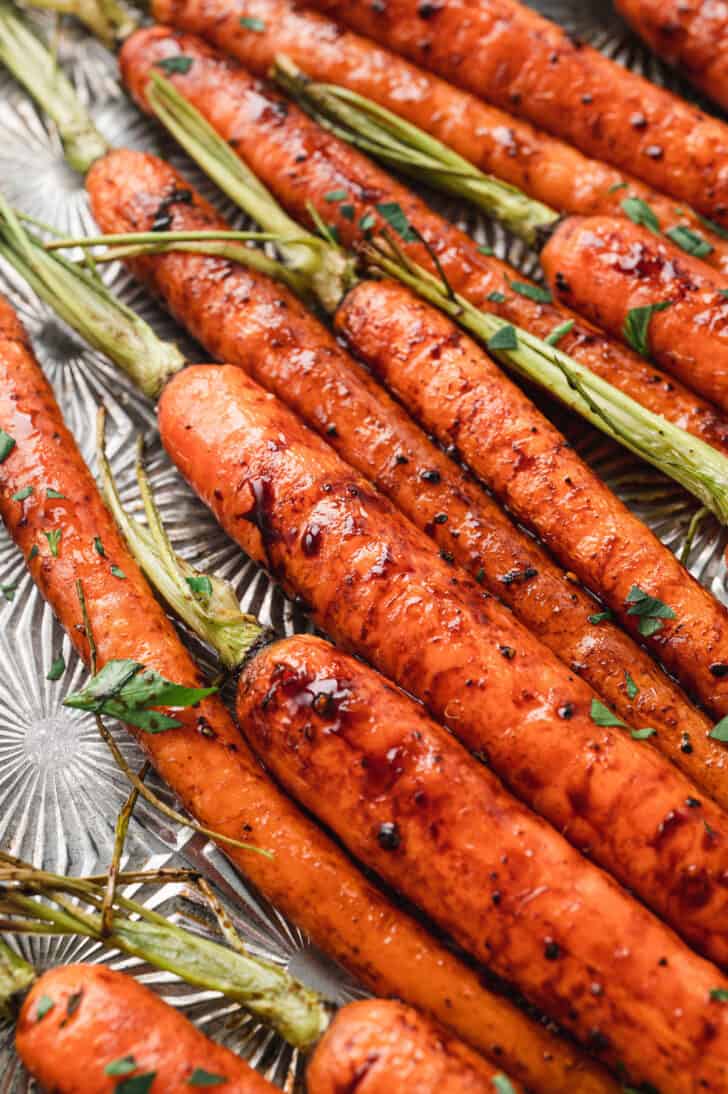 A textured baking pan topped with balsamic roasted carrots with their green tops still on, sprinkled with chopped parsley.