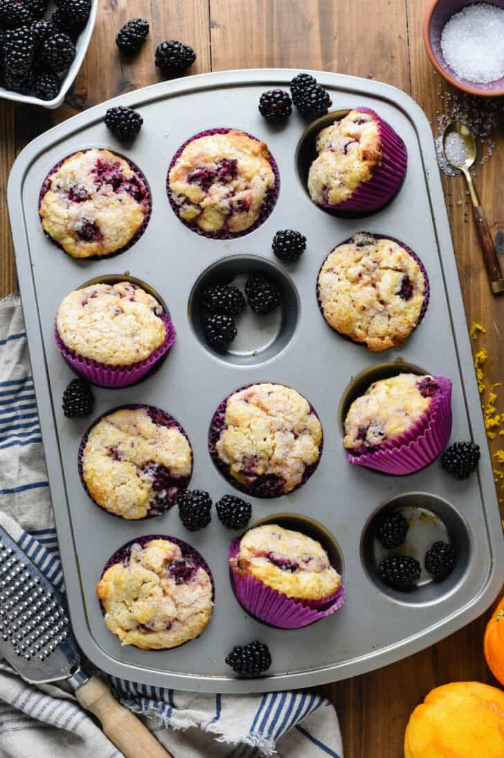 A gray metal baking pan with ten blackberry muffins in purple paper liners, along with fresh blackberry arranged around the muffins.