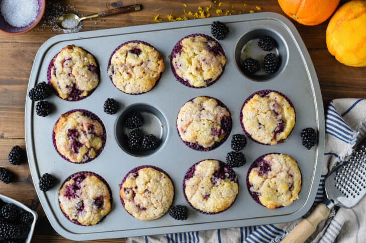A gray metal baking pan with ten muffins with blackberries in purple paper liners, along with fresh blackberry arranged around the muffins.