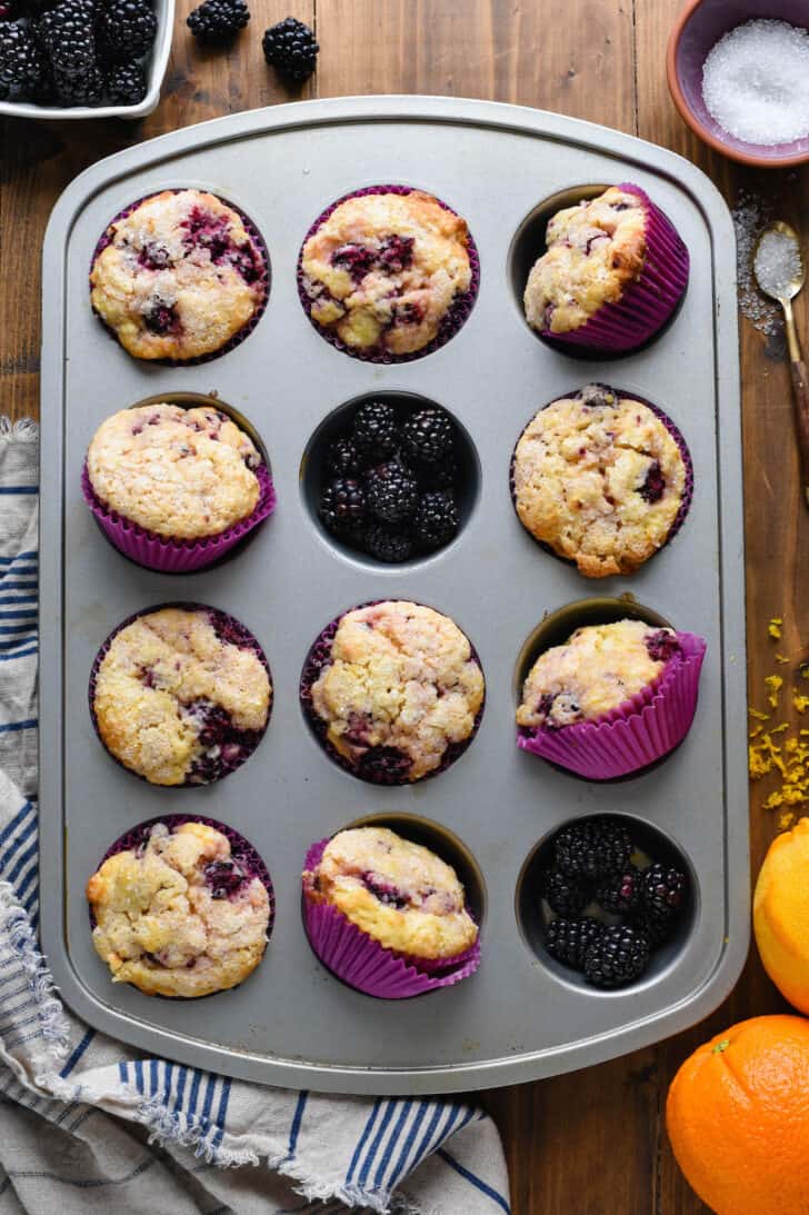 A gray metal baking pan with ten fruit quickbreads in purple paper liners, along with fresh fruit arranged in two cupcake wells.