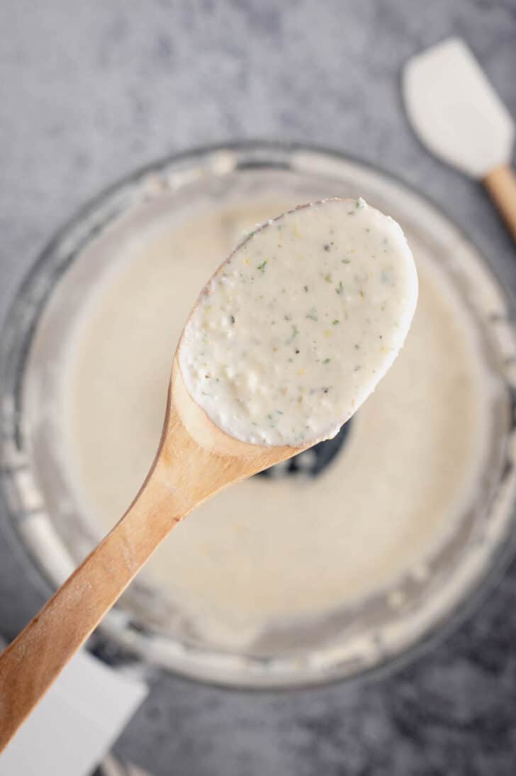 A wooden spoon lifting up a blended white mixture flecked with green herbs.