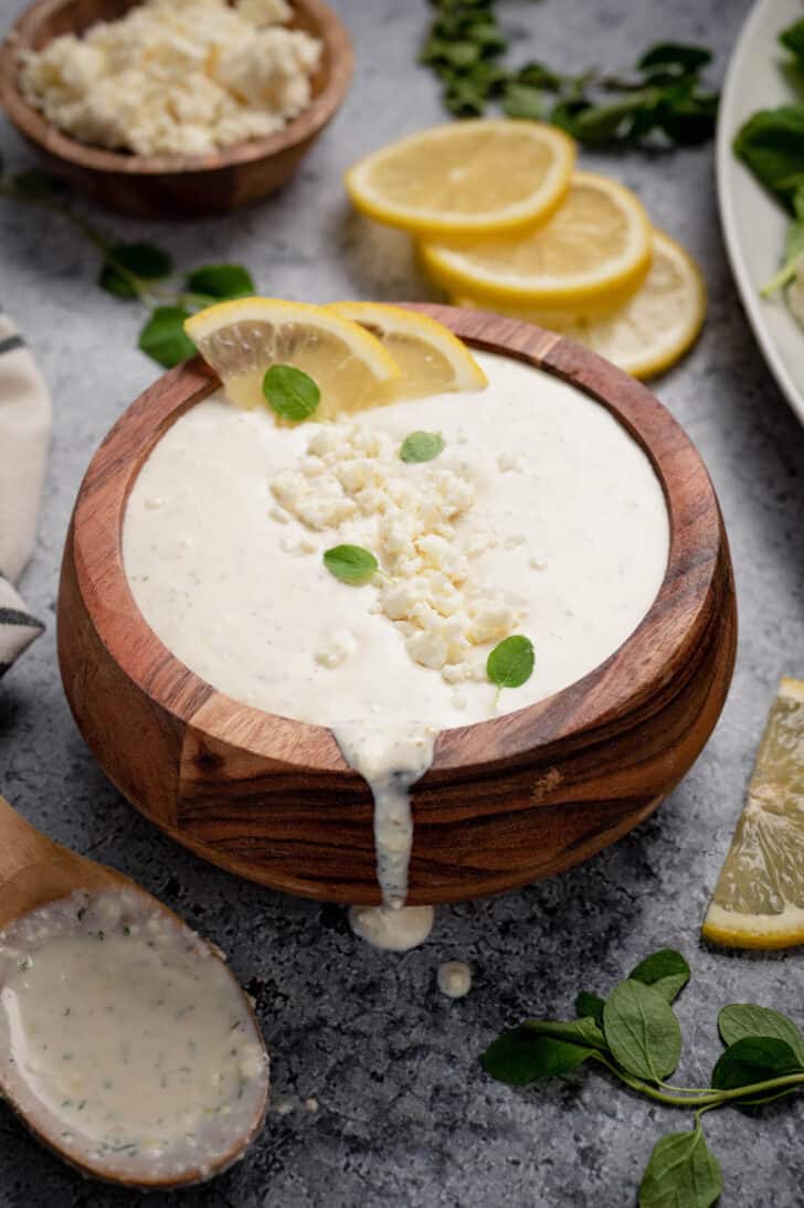 A small wooden bowl filled with a milky cheese sauce, garnished with crumbled white cheese, lemon wedges and herb leaves.