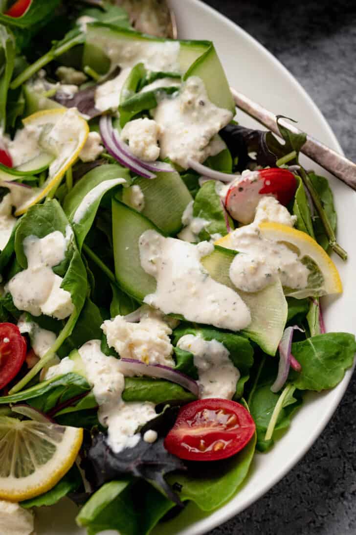 A large white platter filled with salad made from mixed greens, cucumber ribbons and grape tomatoes, tossed with feta dressing.