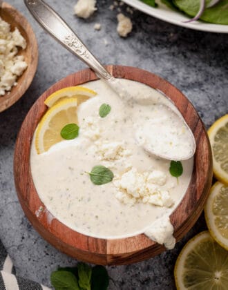 A small wooden bowl filled with creamy feta dressing, garnished with fresh lemon wedges and oregano leaves, with a spoon digging into it.