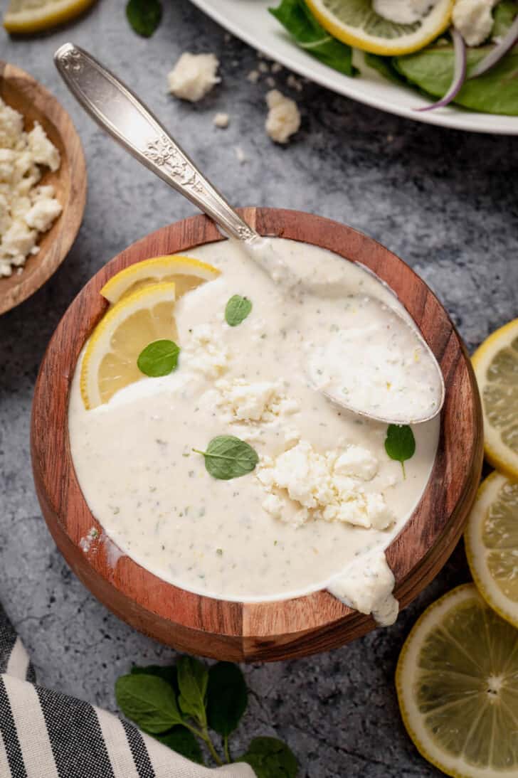 A small wooden bowl filled with creamy feta dressing, garnished with fresh lemon wedges and oregano leaves, with a spoon digging into it.