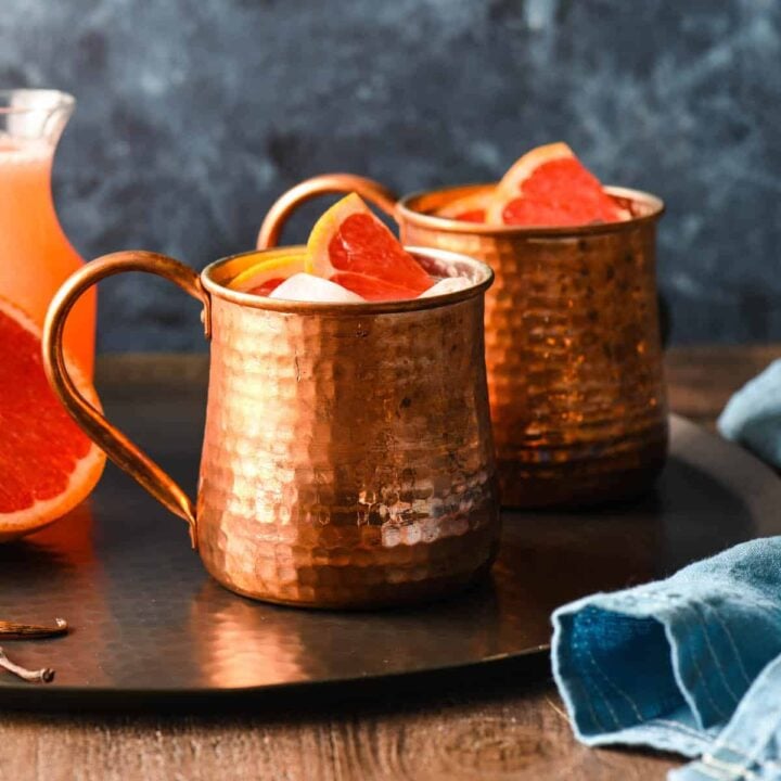 Two grapefruit mules in hammered copper mugs on a black tray with half a grapefruit and a bottle of grapefruit juice.