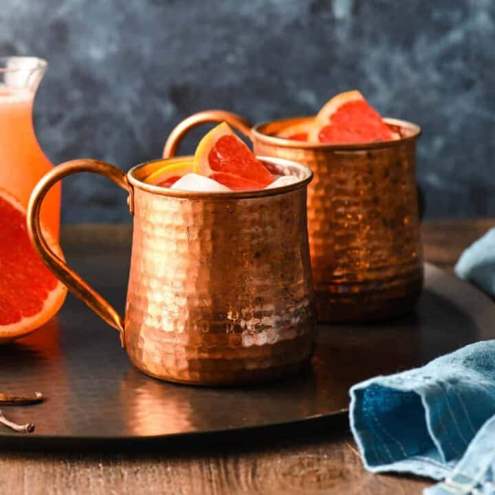 Two grapefruit mules in hammered copper mugs on a black tray with half a grapefruit and a bottle of grapefruit juice.