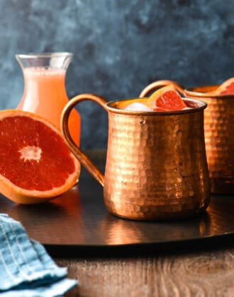 Two grapefruit mules in hammered copper mugs on a black tray with half a grapefruit and a bottle of grapefruit juice.