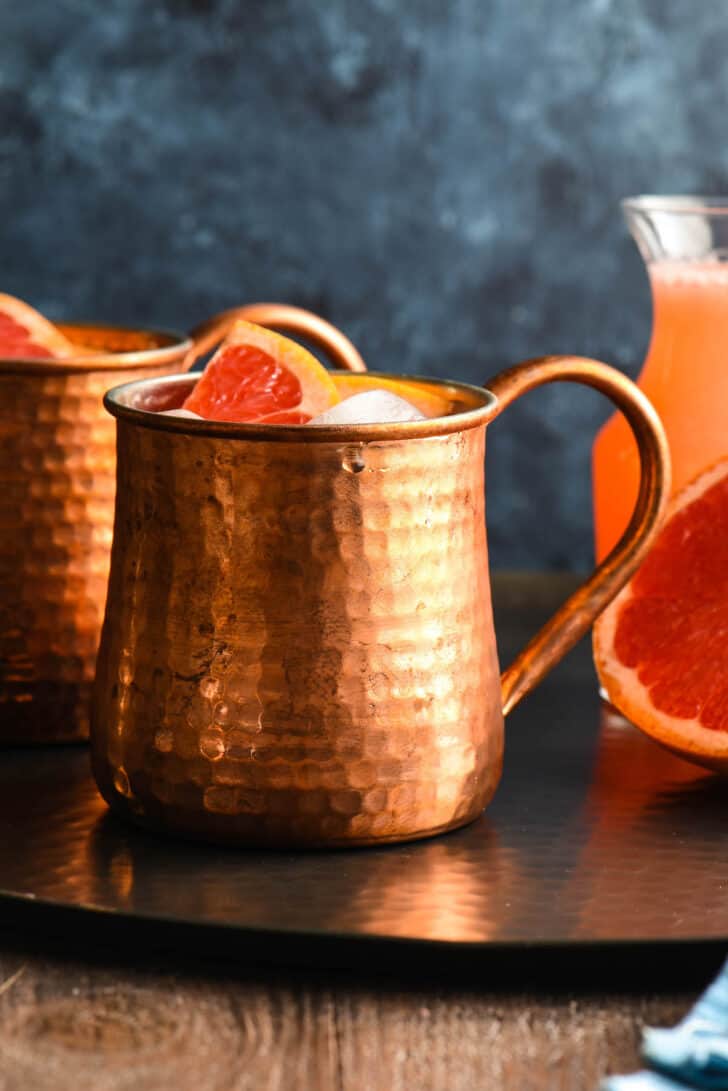 A grapefruit Moscow mule in a hammered copper mug on a black tray with half a grapefruit and a bottle of grapefruit juice.